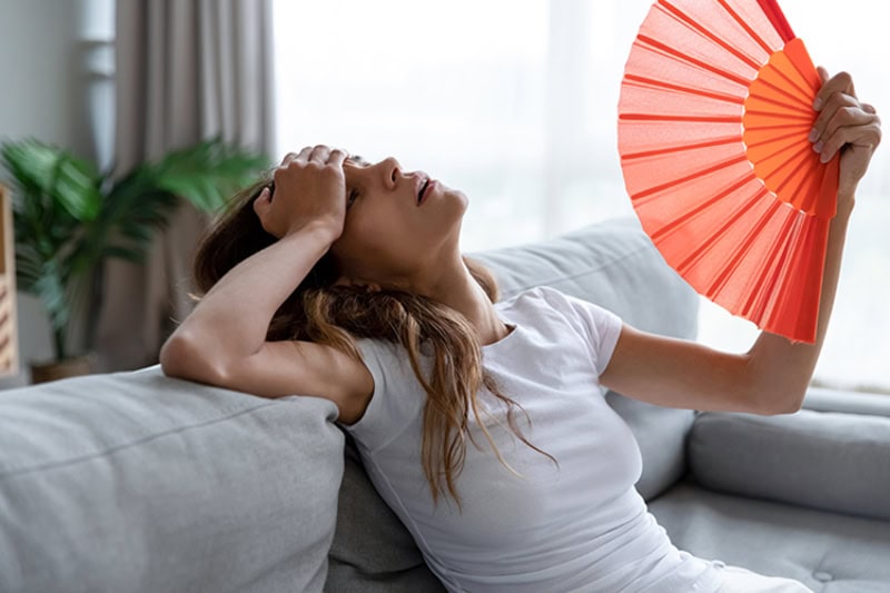 Woman on couch trying to fan herself, because her AC keeps blowing out hot air.