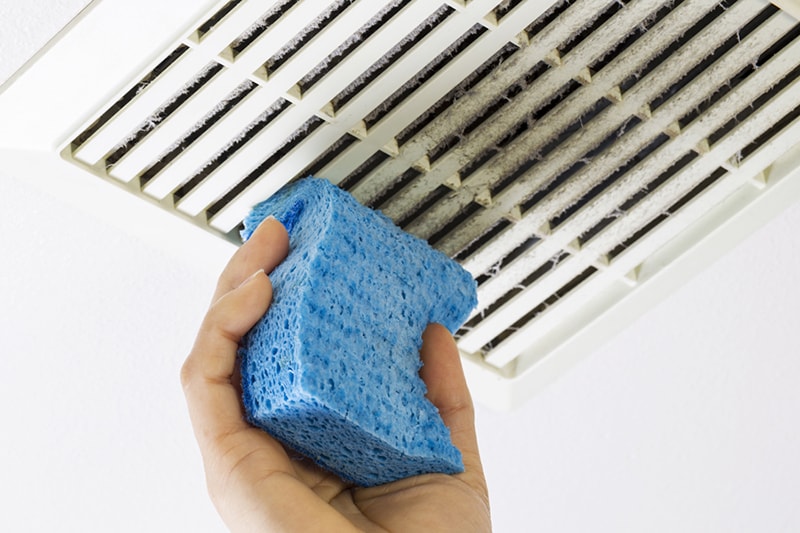 Homeowner cleaning bathroom fan vent with sponge to represent the need for air duct cleaning.