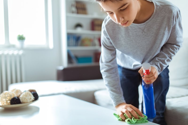 Young child cleaning small table with rag to reduce dust in the home.