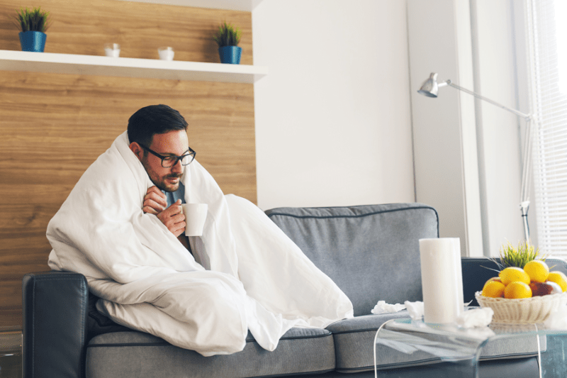 Male homeowner sitting on couch covered in blanket with coffee, because his furnace isn't heating the entire home.
